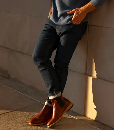 Man wearing blue jeans and brown chelsea boots leaning against wall