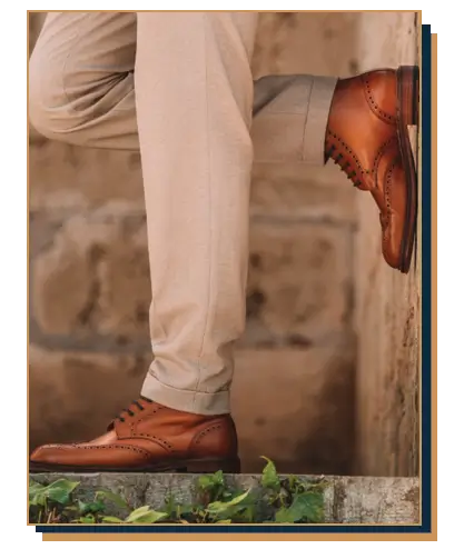 Polished tan leather wingtip brogue dress boots paired with beige cuffed trousers against a stone wall