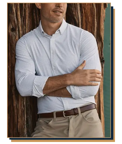 Man leaning against a wooden post wearing a white grid-check button-up shirt with rolled sleeves from Mizzen and Main, tucked into khaki pants with a brown leather belt