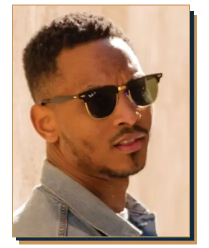 Man wearing a light wash denim jacket, jeans, and tortoiseshell browline sunglasses leaning against a stone wall outdoors